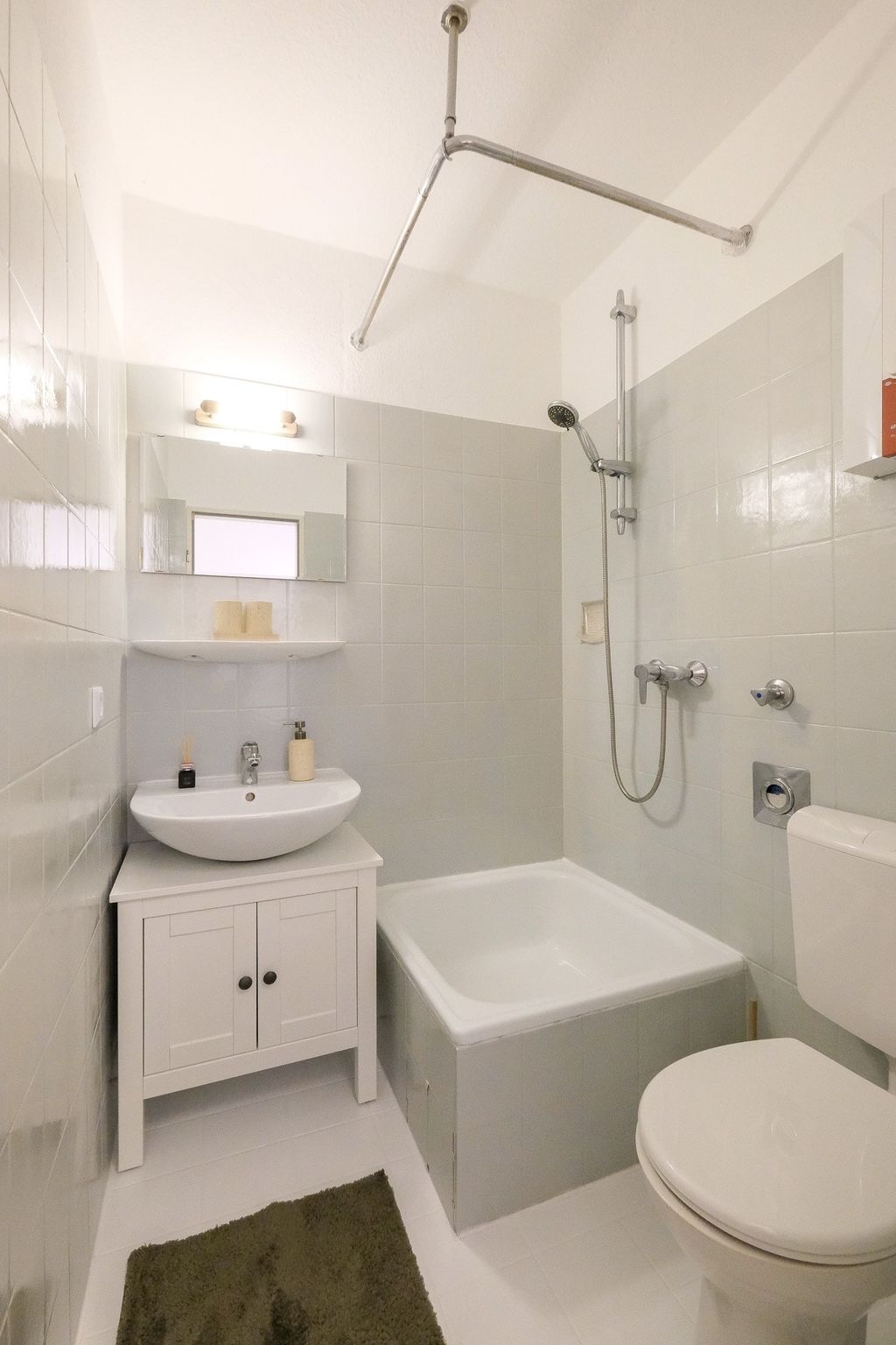 Small bathroom with sink on a white cabinet, toilet, and bathtub with showerhead, gray tiles, and a dark green bath mat on the floor.