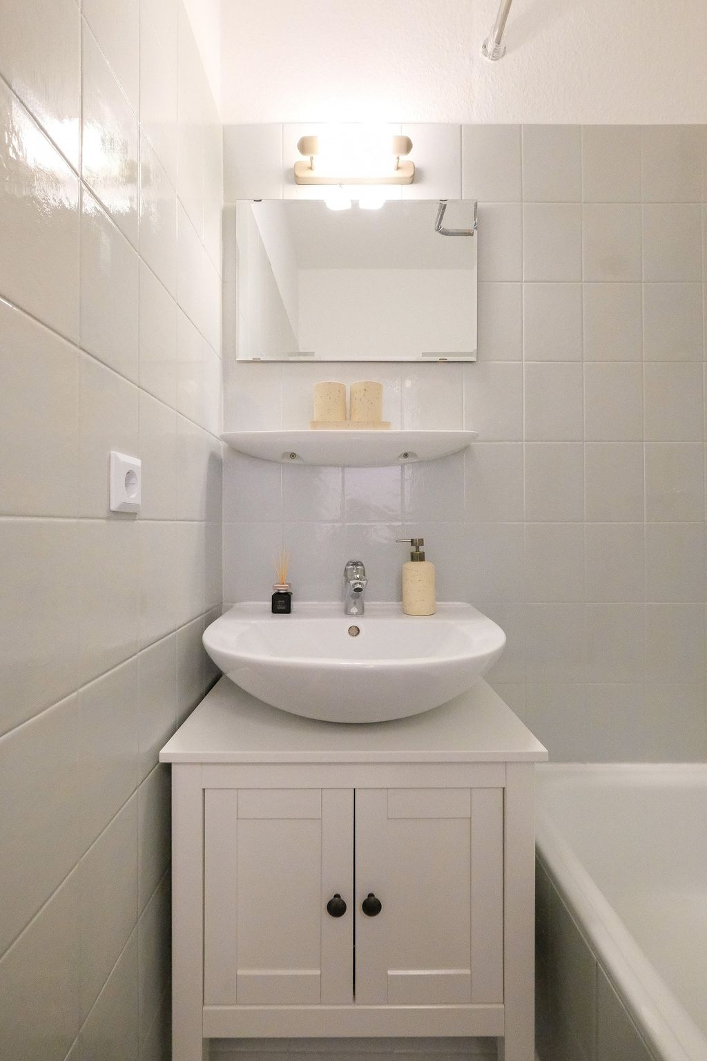 Small bathroom with a white sink on a white cabinet, a mirror above, and two candles on a floating shelf, surrounded by beige-white tiles.