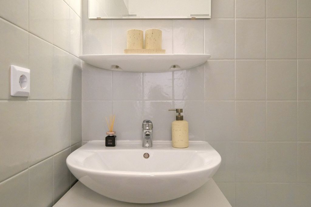 White sink with chrome faucet in a bathroom with light gray tiled walls, featuring a soap dispenser, a reed diffuser, and two candles on a shelf below a mirror.