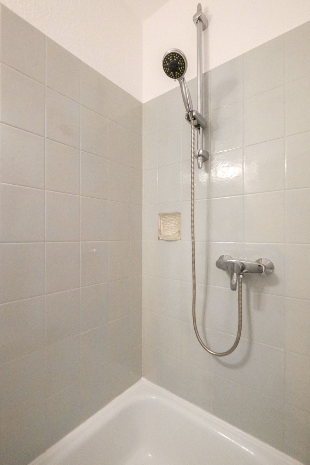 A simple shower area with white wall tiles, an adjustable handheld showerhead, and a faucet above a bathtub.