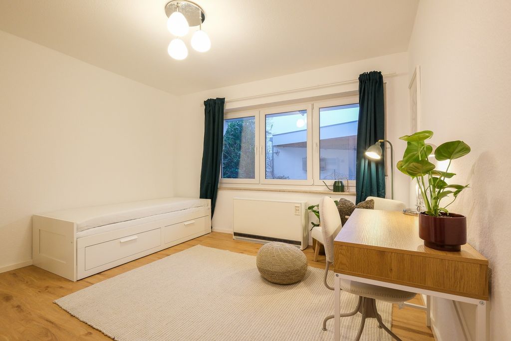 Brightly furnished room featuring a daybed with drawers, a desk with potted plants, a chair, and a large window with dark curtains.