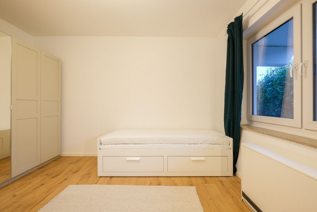 A small, simply furnished room featuring a white single bed with two drawers, a gray curtain beside a window, and a large wardrobe with a mirror on the wall.