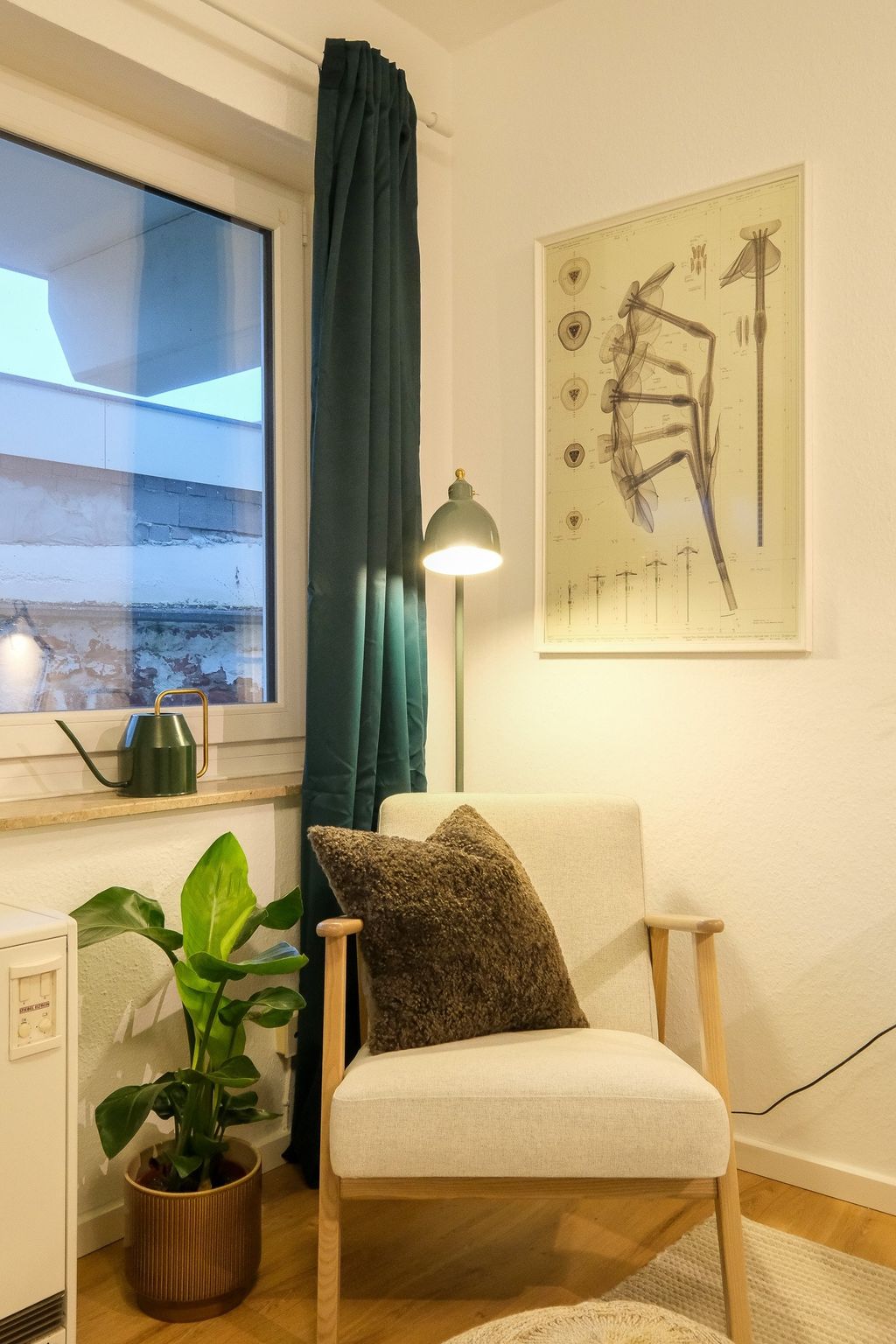 A cozy room featuring a beige armchair with a brown cushion, next to a potted plant and a window with blue curtains, along with a green floor lamp and a botanical poster on the wall.