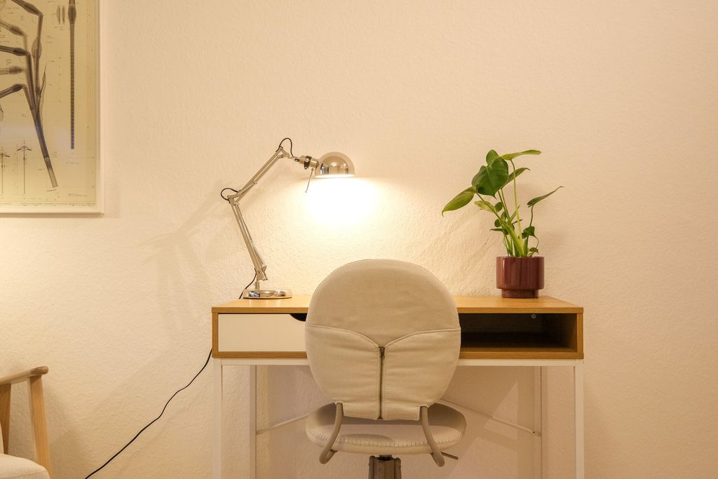A simple desk with an adjustable desk lamp, a potted plant, and a beige office chair in front of a white wall.