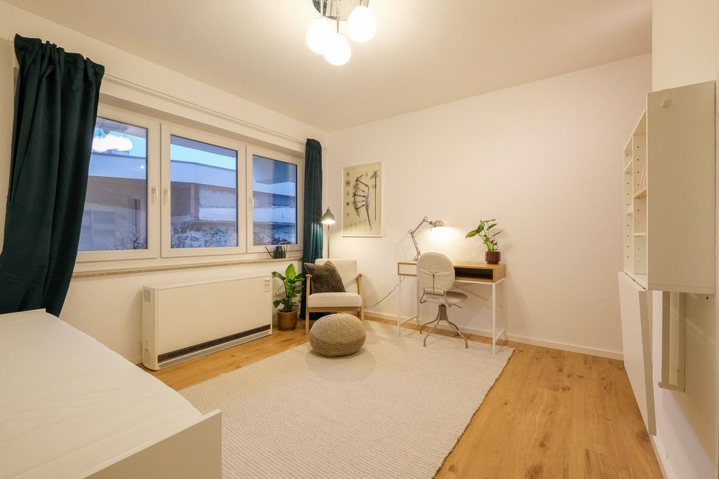 A cozy room with light walls, wood flooring, a light-colored desk with a chair and lamp, an armchair with a pillow, a plant pot, large windows with dark green curtains, and a light carpet on the floor.