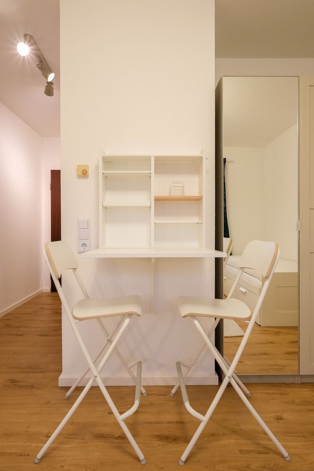 Small dining area with white folding chairs and a wall-mounted white folding table in a modern, bright room with wooden flooring.
