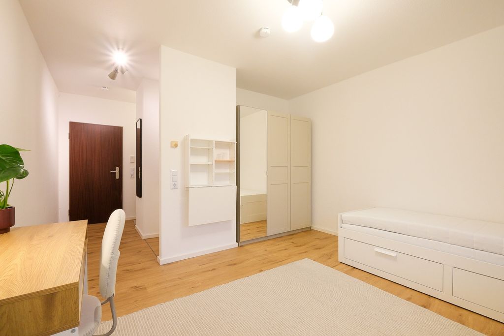 Bright, minimally furnished room with wooden floor, a desk with chair on the left and a storage bed on the right, white walls, and a large mirror on a wardrobe.