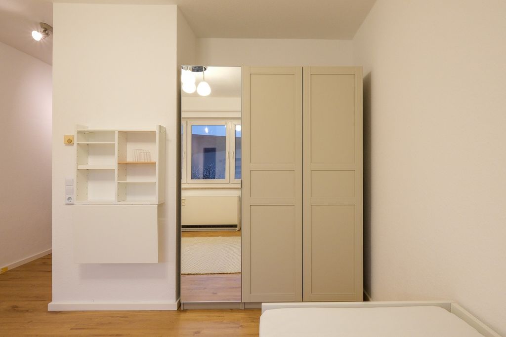 A bedroom featuring a white double wardrobe with one mirrored door. Open wall shelves are mounted on the left wall, and part of a bed is visible in the foreground.