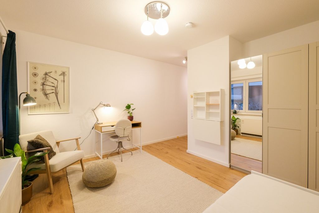 Bright, minimalist workspace featuring a white desk, chair, armchair, and a large wall mirror, with wood flooring and a cream rug.
