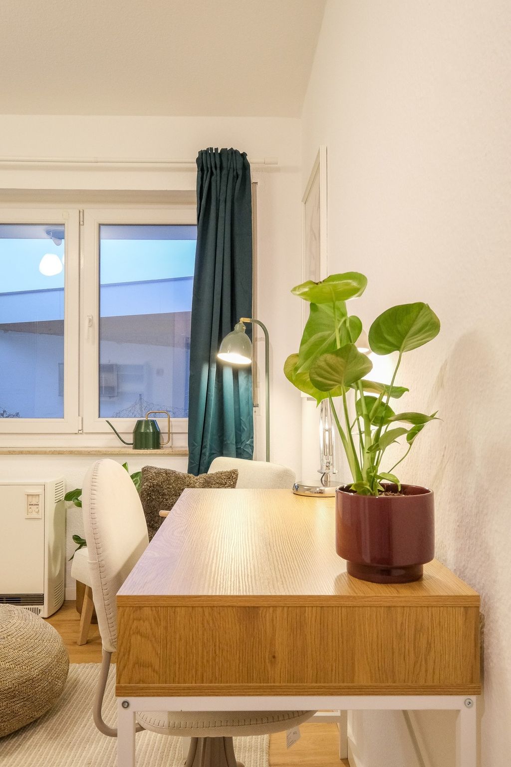 Modern light wood desk against a white wall with a potted plant, table lamp, and a window with dark blue curtain in the background.