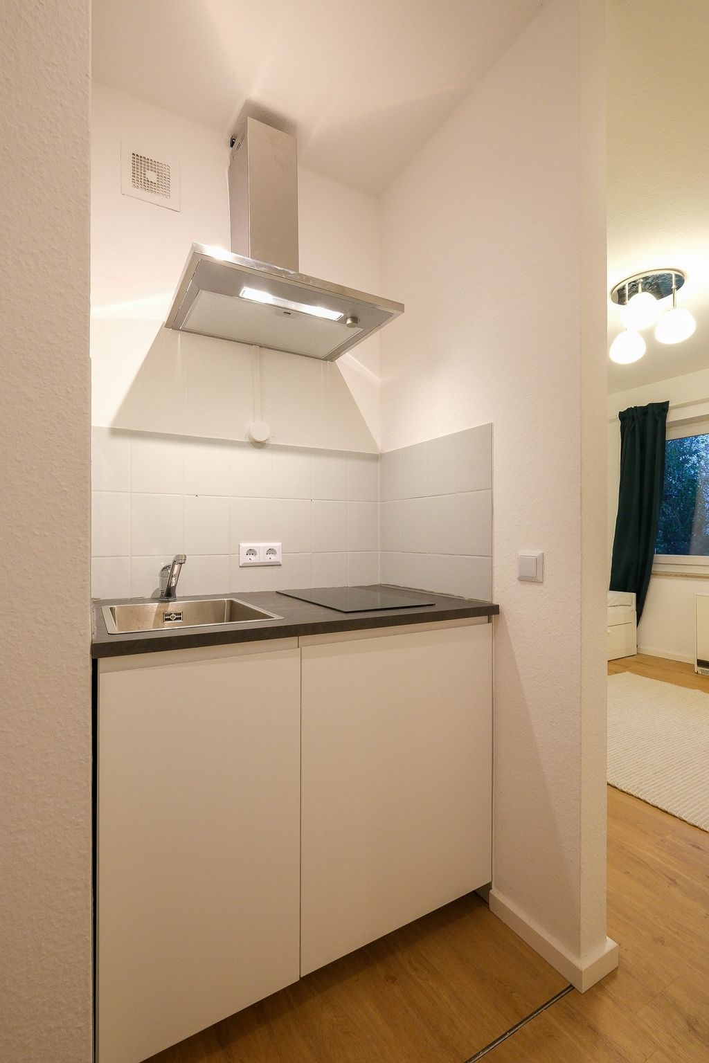 Small, modern kitchenette with a sink, induction cooktop, and stainless steel extractor hood in a white room with wooden flooring.