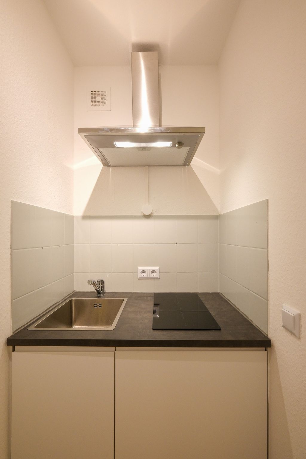 Small modern kitchen with sink, cooktop, and stainless steel extractor hood in a niche with white tiles.