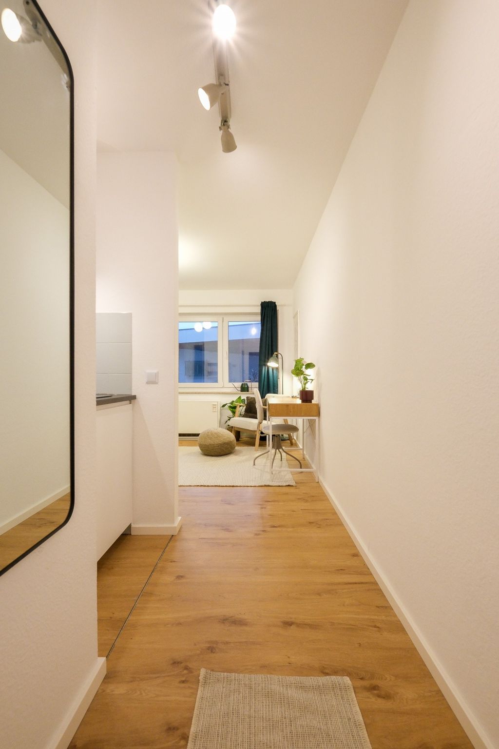 A bright hallway with wooden flooring leads to a cozy area with armchairs, a pouf, a desk with a lamp, and plants in front of a window with dark green curtains.
