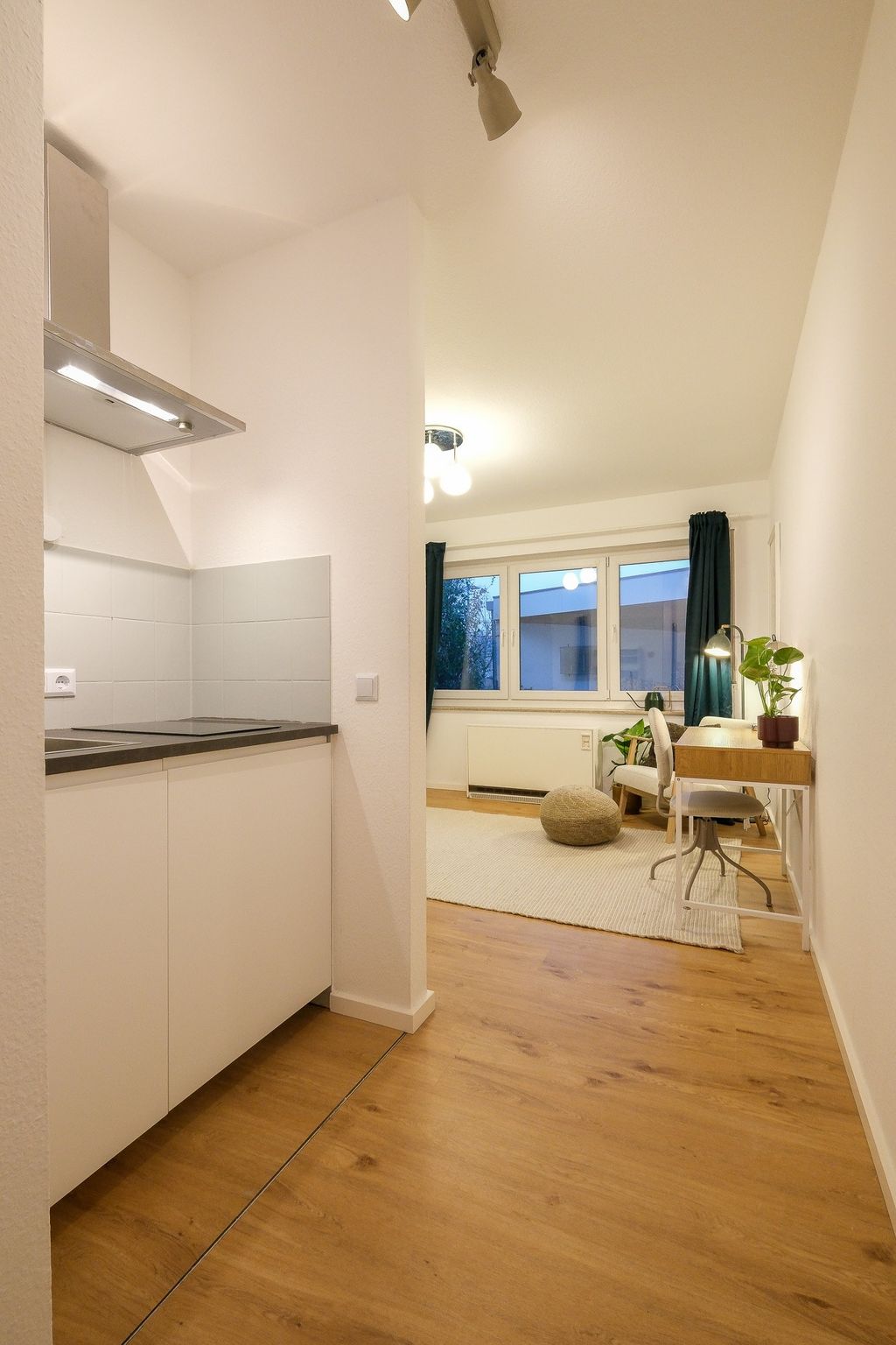 A bright room with laminate flooring, a small white kitchenette on the left, and a workspace with a desk, chair, plants, and a window with blue curtains in the background.