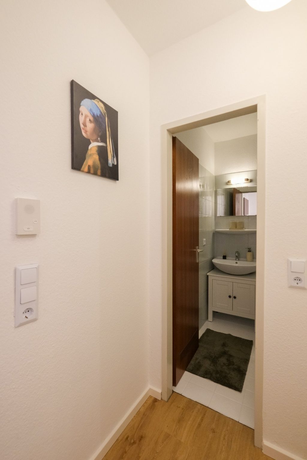 View into a small washroom with an open wooden door, white cabinet, and gray bath mat, with a painting of the Girl with a Pearl Earring on the wall to the right.