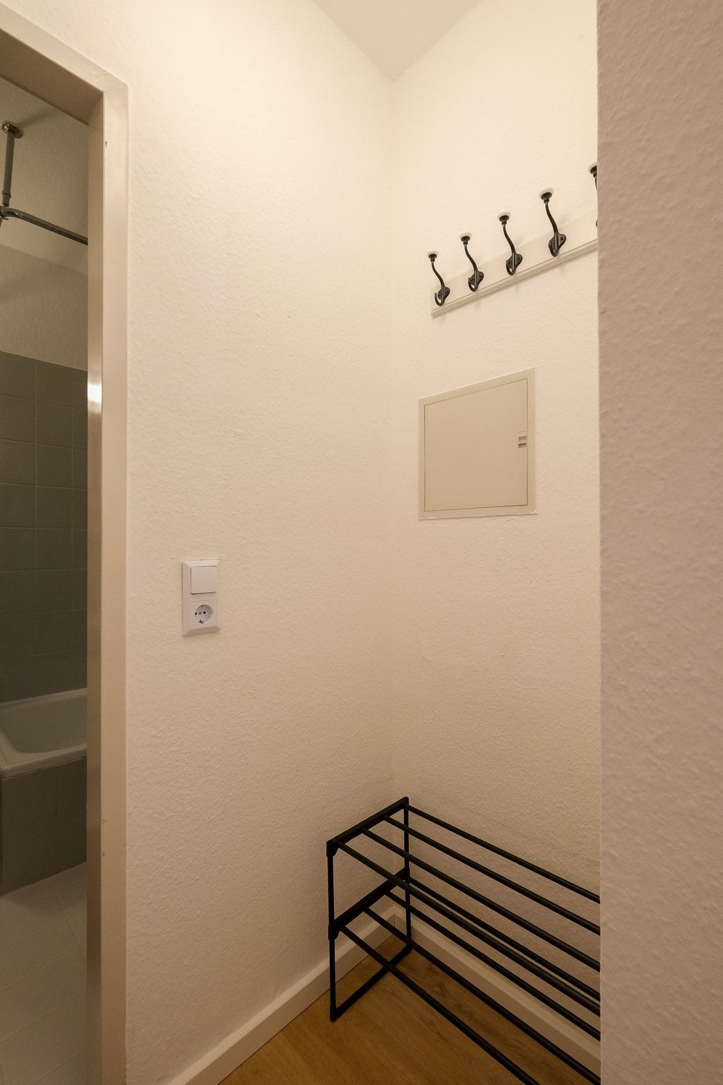 A small hallway corner with a black metal shoe rack, four black wall hooks, and an open door leading to a bathroom with gray tiles.