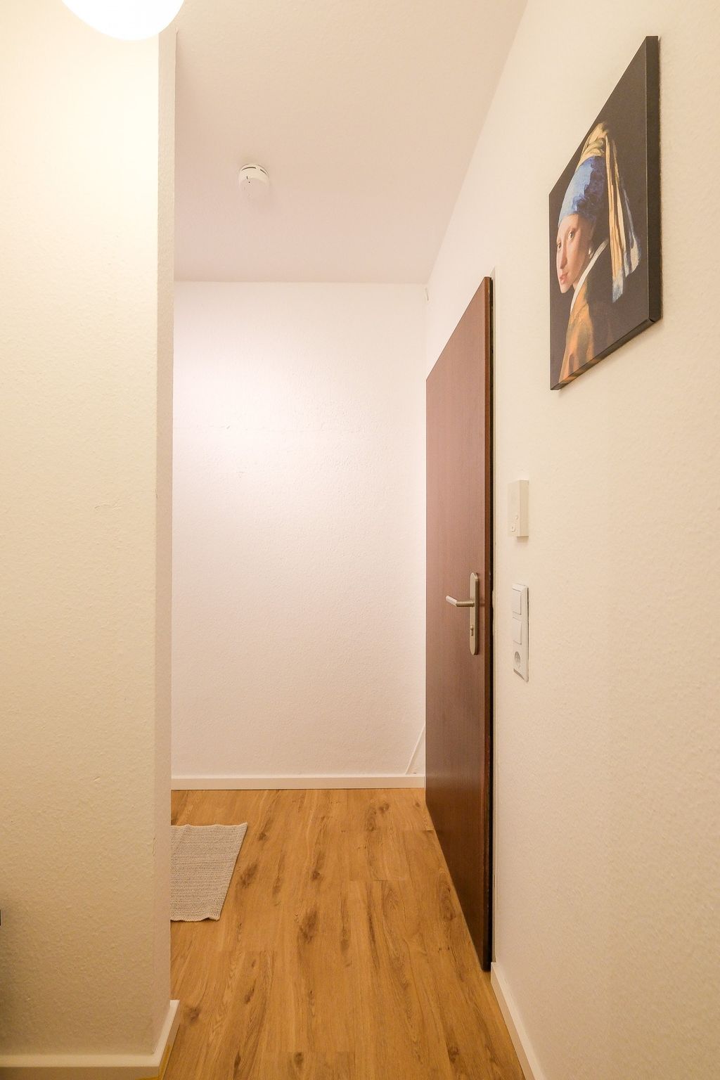 A bright hallway with an open wooden door, a white light switch, and a painting of the Girl with a Pearl Earring on the wall.