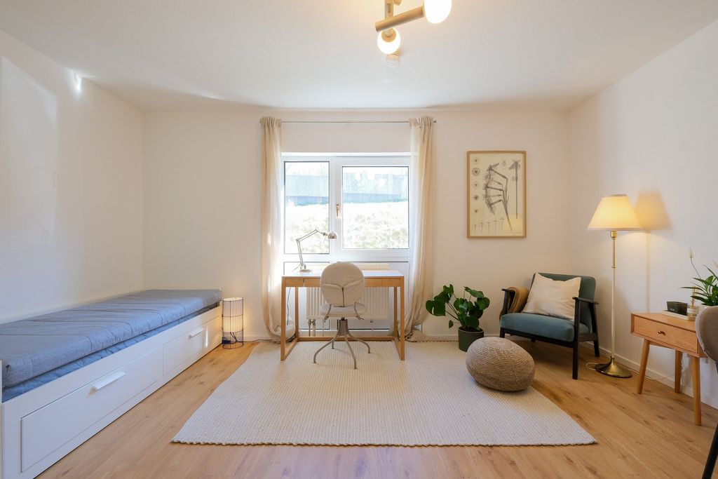 Bright, minimalist office room with wooden floor, white desk in front of a window, gray swivel chair, a daybed on the left, and a cozy armchair with a floor lamp on the right.