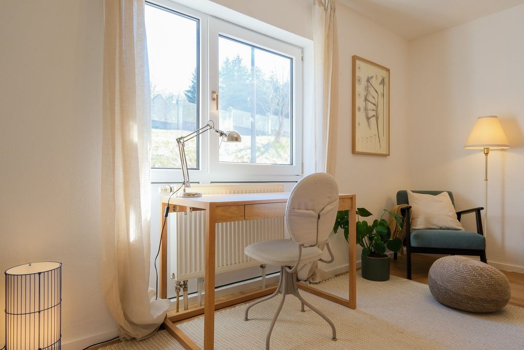 Bright workspace featuring a simple wooden desk in front of a window, a grey office chair, a green plant, a comfortable armchair with a cushion, and two floor lamps.