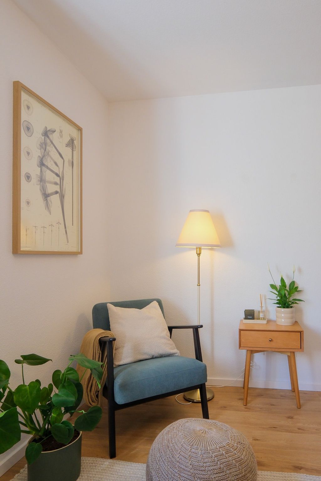 A cozy reading nook with a blue armchair featuring a cushion and a blanket, next to a small wooden side table with a plant and decorative items, plus a floor lamp.