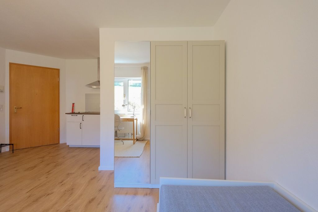 A bright room with wooden flooring featuring a white wardrobe with a mirror, partially showing a kitchen area and a wooden door.