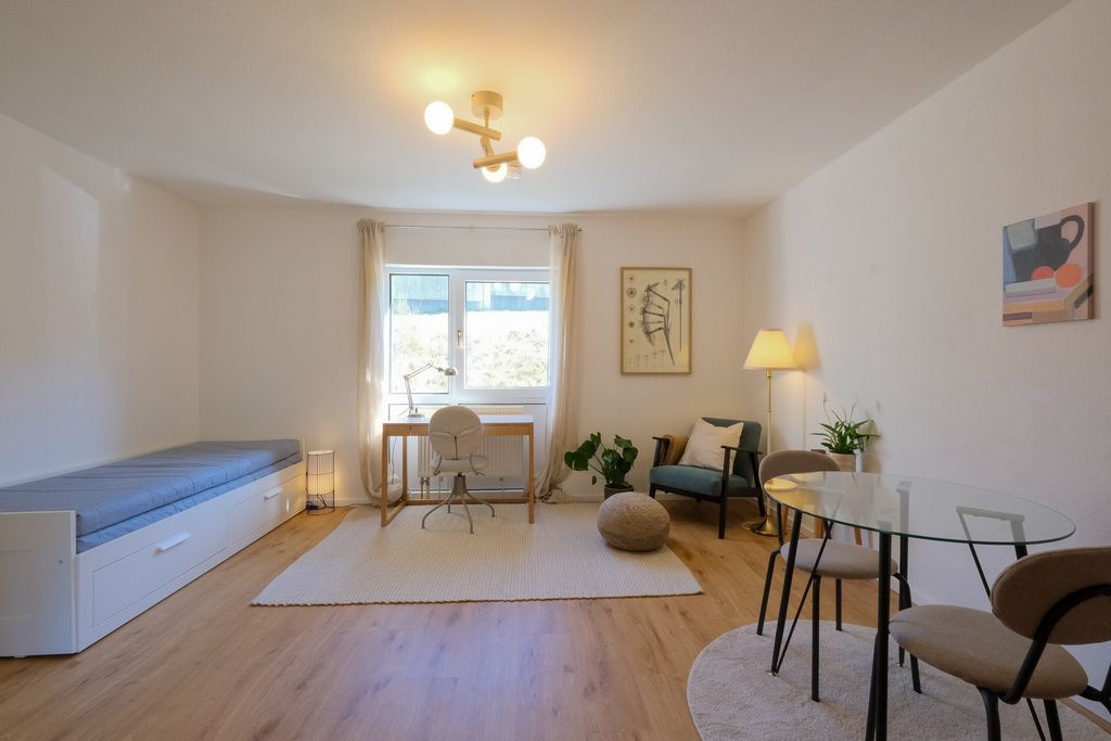Bright modern living room with a sofa, a desk in front of a window, a chair, a round glass table with chairs, and a floor lamp.