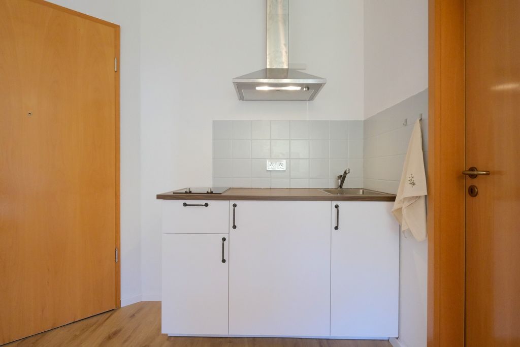 Small modern kitchenette with white cabinet fronts, brown countertop, stainless steel range hood, built-in cooktop, and sink, flanked by two wooden doors on either side.