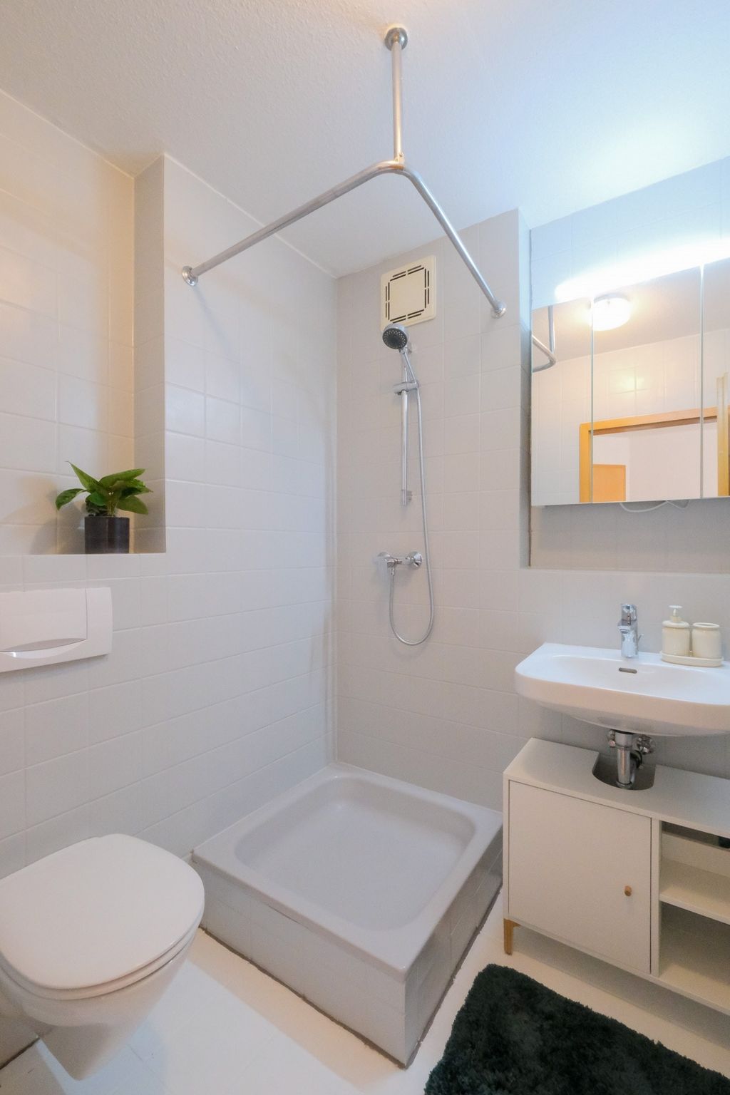 Small, minimalist bathroom with a shower area, white tiled walls, toilet, sink with cabinet, mirror, and a small potted plant on a wall niche.