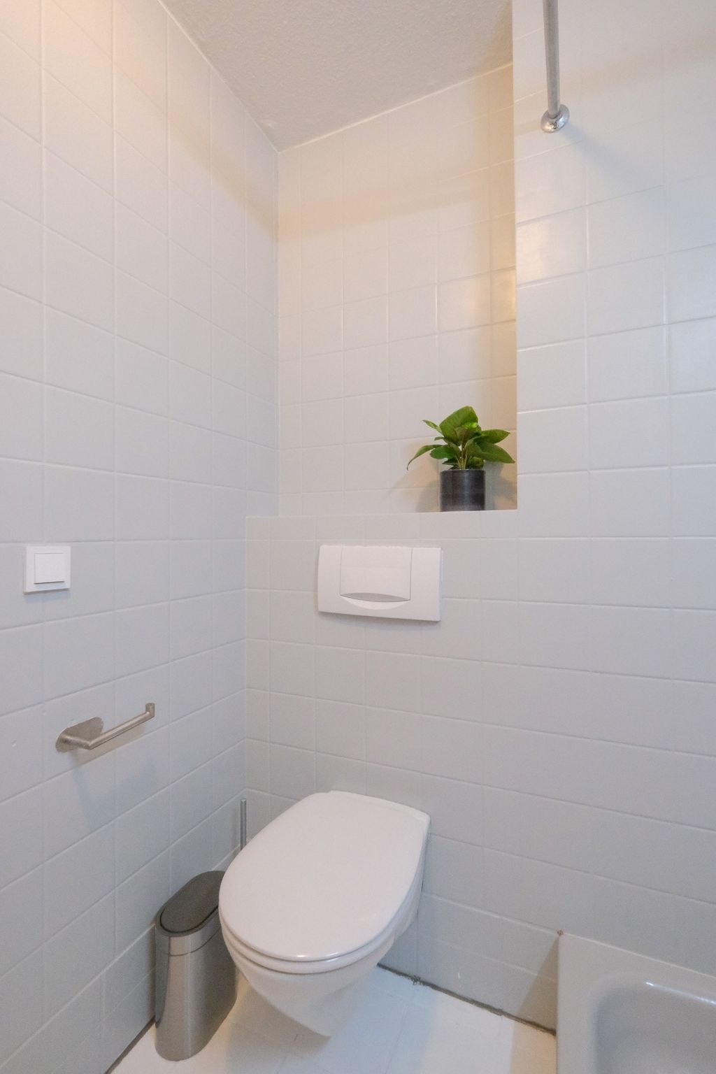 Small modern bathroom with white tiled walls, wall-mounted toilet, metal trash can, and a small green potted plant in a wall niche.