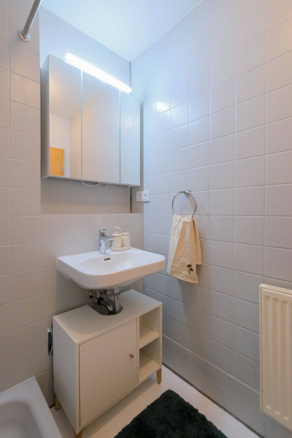 Small bathroom featuring a white sink mounted on a white cabinet, a wall mirror with light above, and a towel hanging on a towel ring attached to the tiled wall.