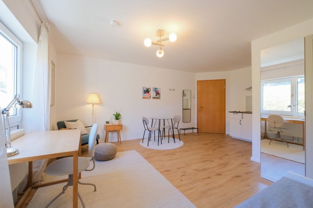Brightly furnished living room with wooden flooring, a round dining table with four chairs, and a workspace with desk and chair by the window.