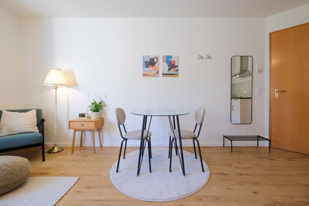 A bright dining area featuring a round glass table with two chairs on a round beige rug against a white wall with two abstract paintings. To the left, a floor lamp stands next to a small wooden table with a plant; to the right, a wooden door and a wall mirror with a shelf are visible.