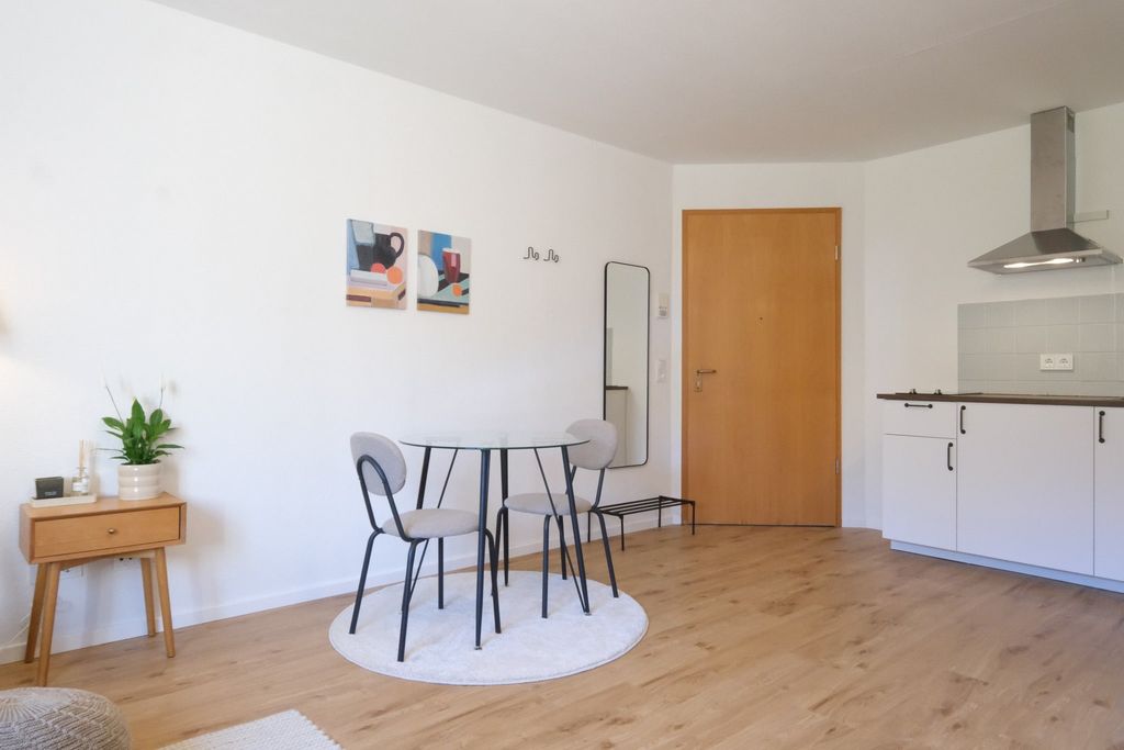 A bright living space with a small round dining area in the center, featuring a white round table and three gray chairs on a round rug. In the background, there is a wooden door, a wall mirror, and a minimalist kitchen unit with a range hood.