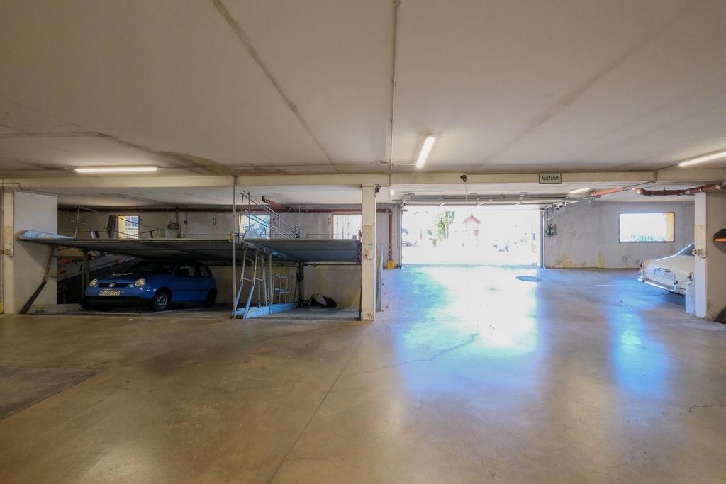 A spacious, empty parking area inside a garage with two parked cars, one blue on the left and one white on the right, and a partially open garage door in the background.