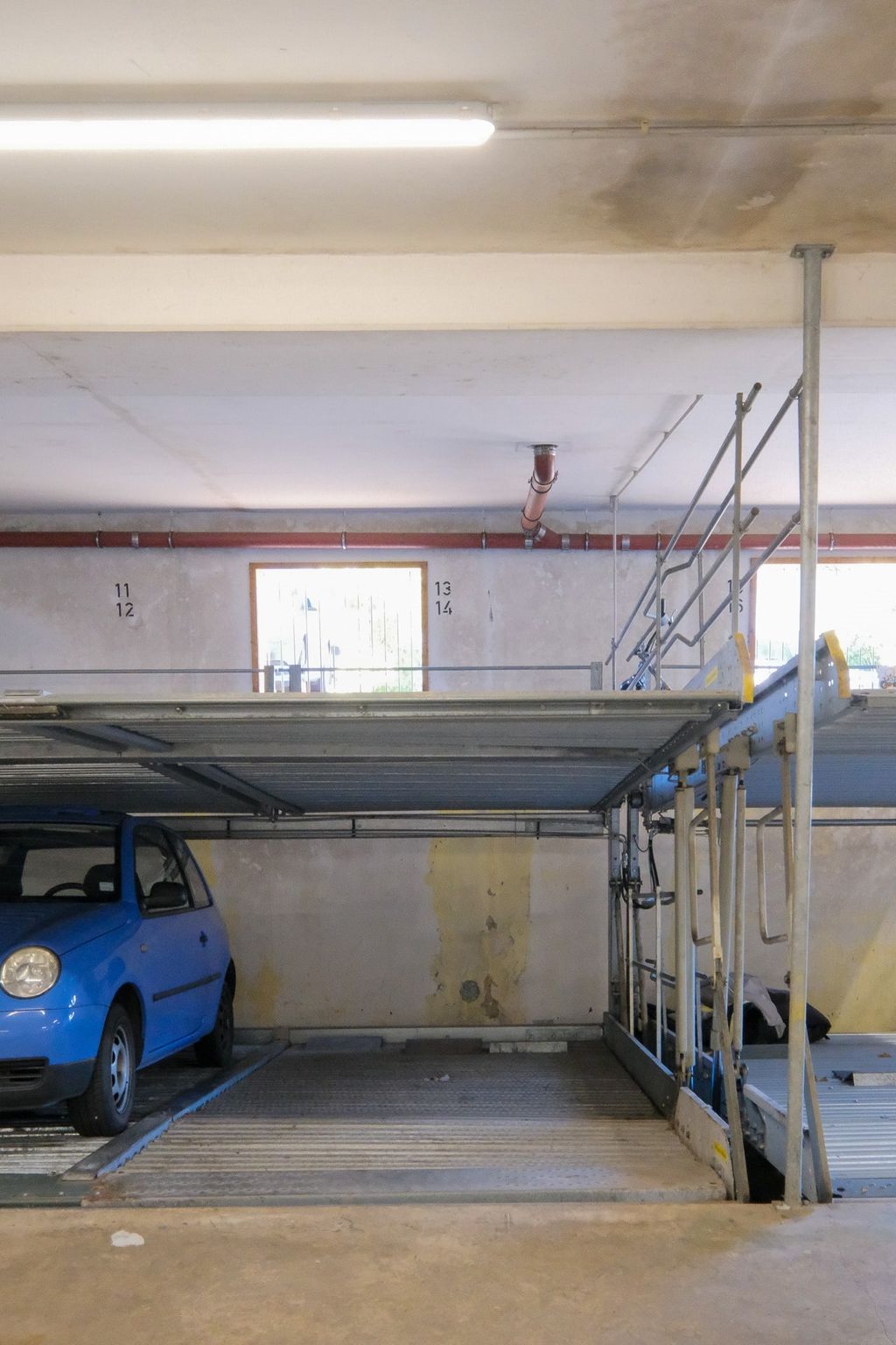 Blue car in a multi-level parking structure with car lift platforms and a window in the background.