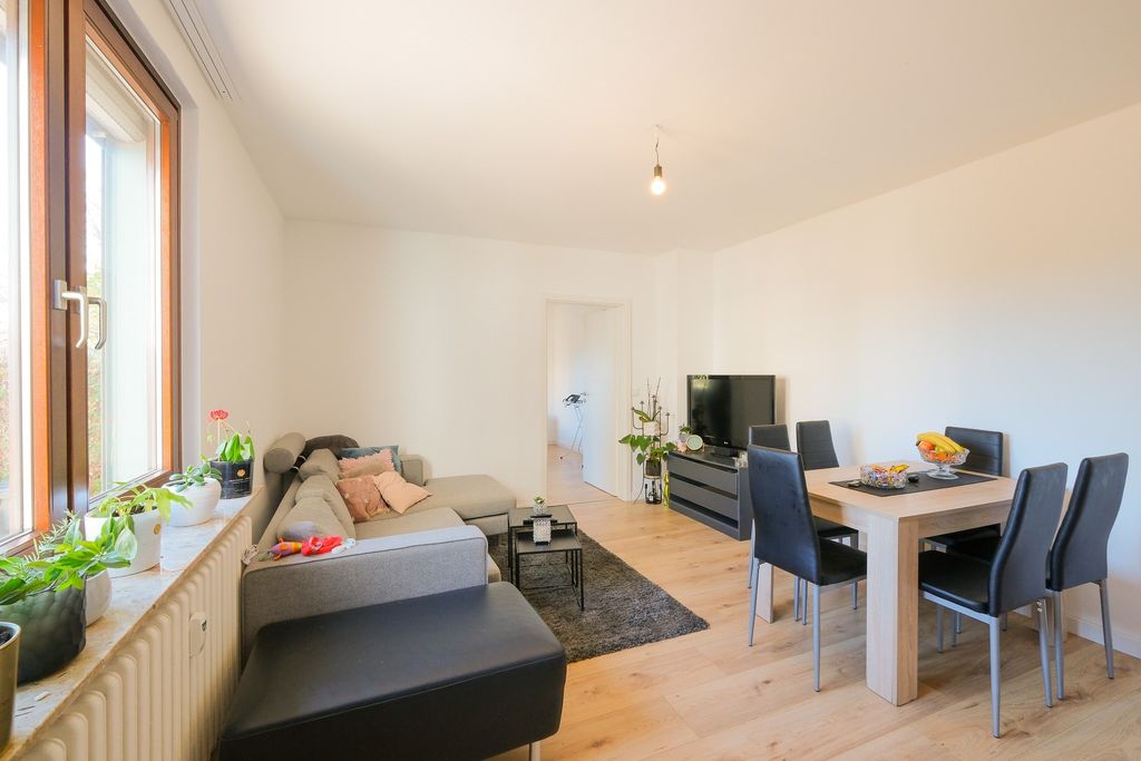 A bright living room featuring a gray sectional sofa, a black ottoman, a dining table with six black chairs, and a flat-screen TV on a sideboard, illuminated by a large window on the left.