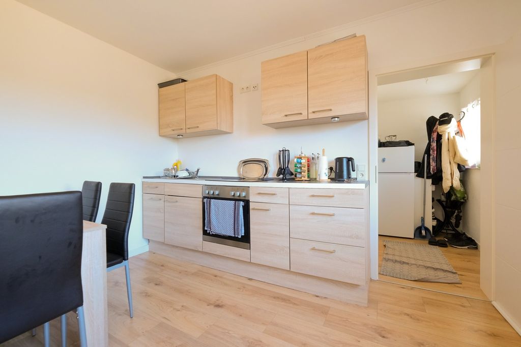 Bright modern kitchen with wooden cabinets, built-in oven, and an adjacent small hallway with coat rack and refrigerator.