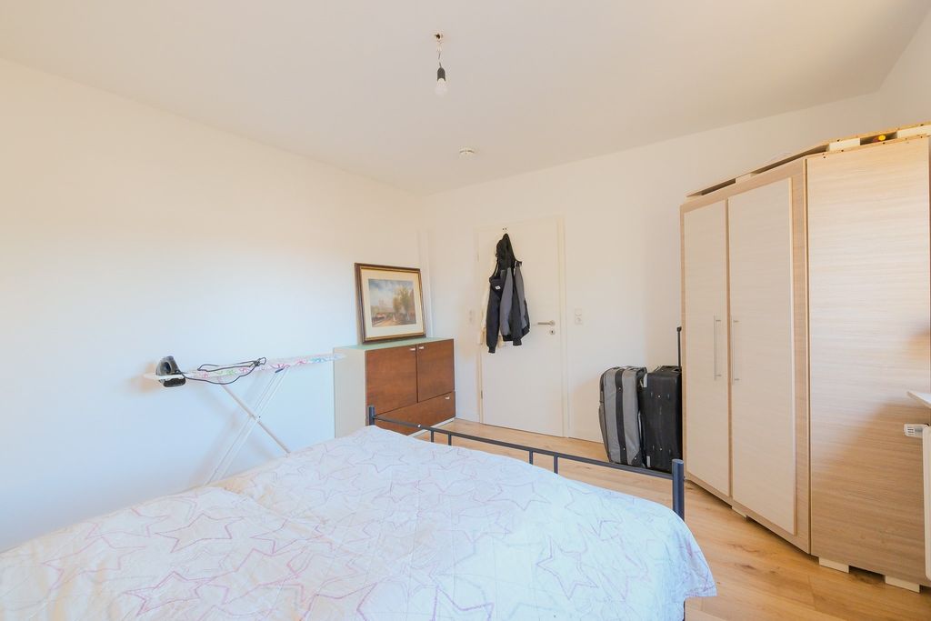 Bedroom with a bed covered by a white blanket, a wardrobe, a chest of drawers with a framed picture, an ironing board mounted on the wall, and two suitcases next to the door with two jackets hanging.