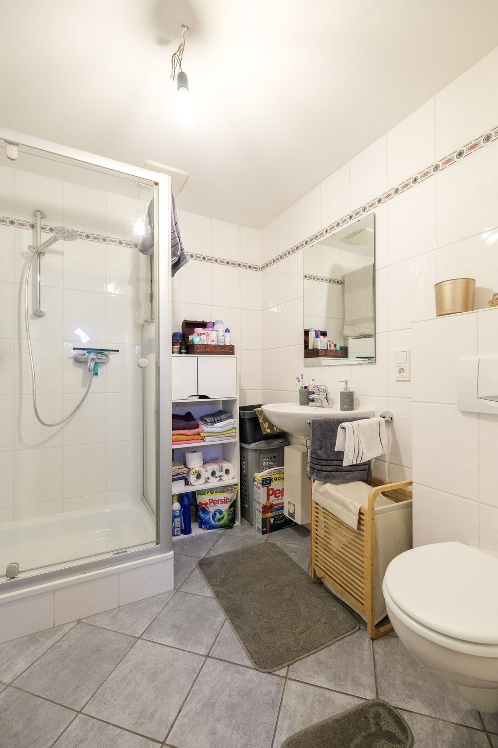 Small white-tiled bathroom with a shower, sink, toilet, mirror, and a storage shelf holding towels and laundry detergents.