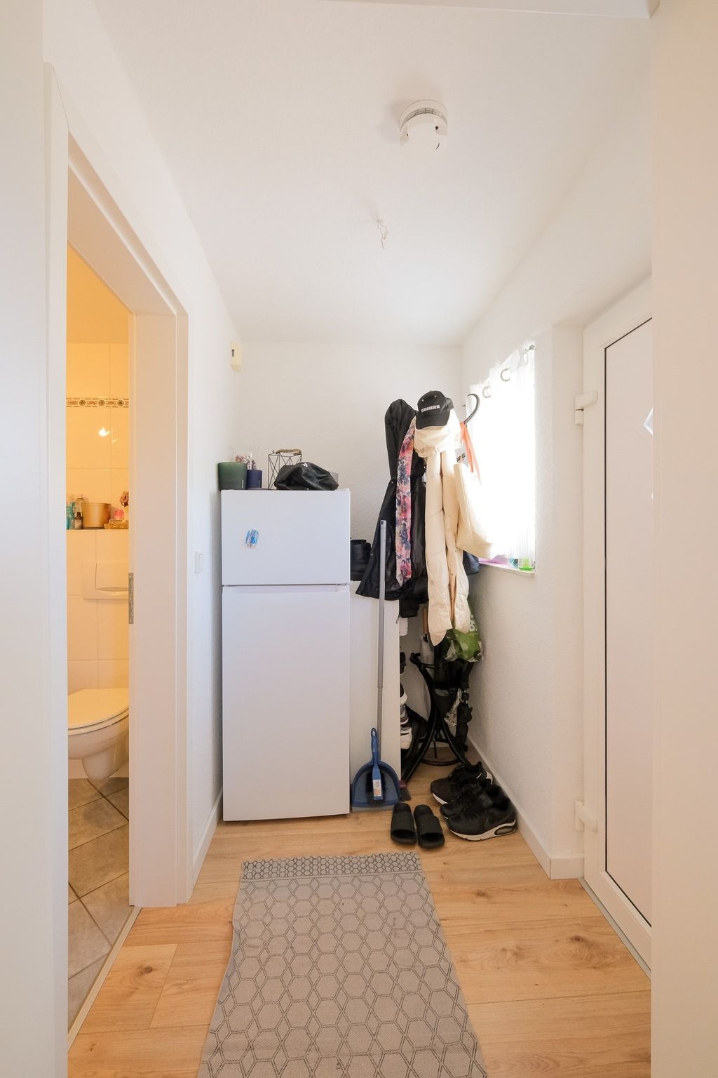 Narrow hallway with a white refrigerator, coat rack with jackets and shoes, and access to a brightly lit bathroom.