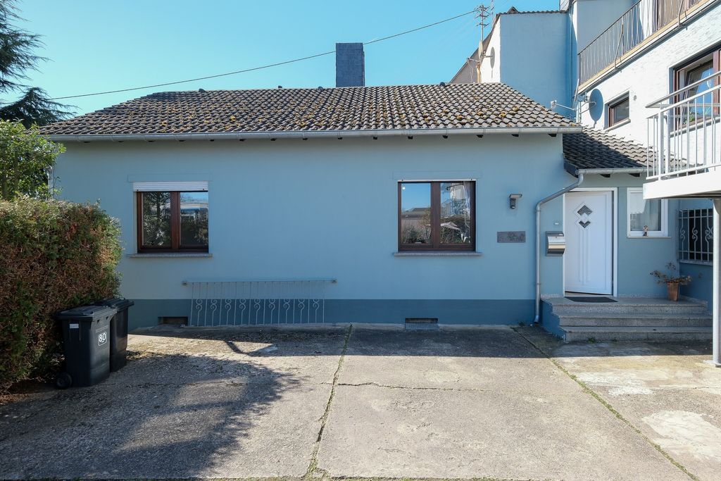 Single-family house with light blue facade, two windows, a white front door on the right, and garbage bins on the left on a concrete yard under a blue sky.