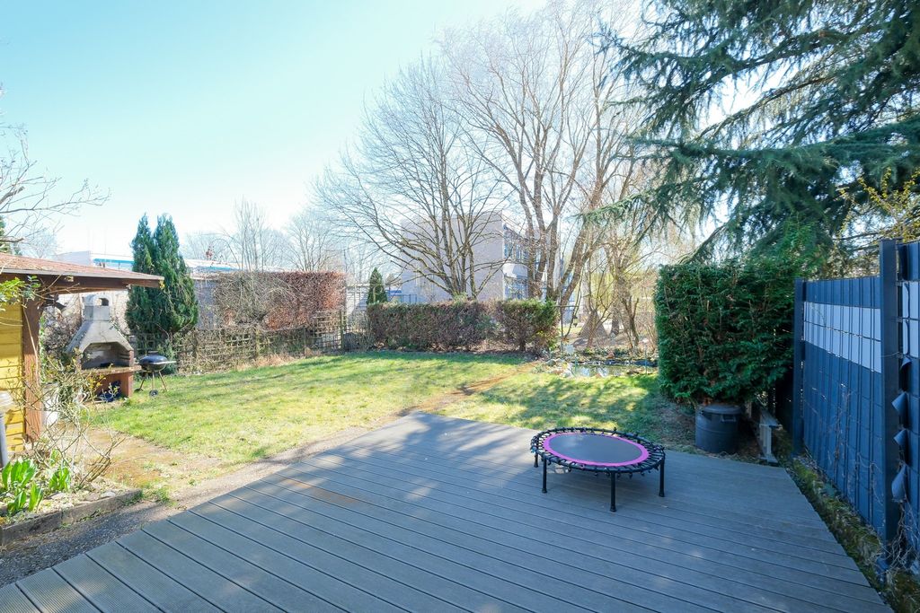A sunny garden featuring a wooden deck with a small trampoline, tall trees, and a barbecue area in the left corner.