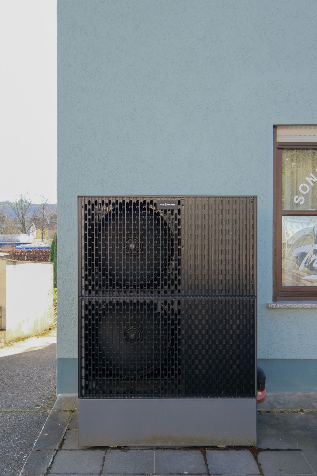 A black Viessmann heat pump is placed in front of a light blue house wall beside a window with curtains.
