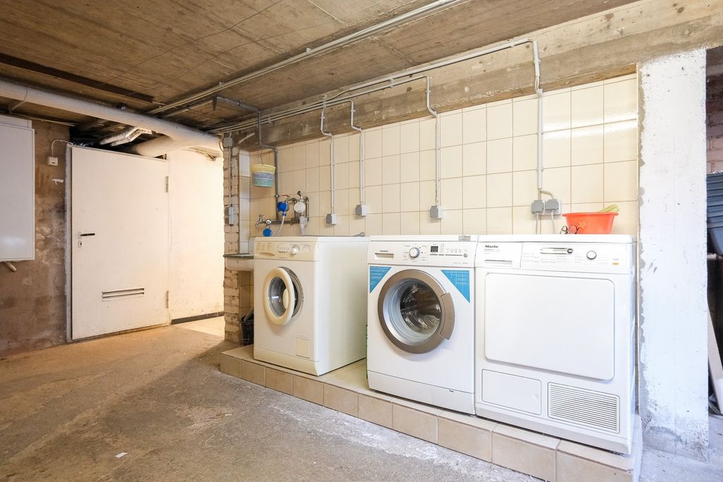 A bare basement room with exposed concrete ceiling, white tiled walls, and three white household appliances (two washing machines and one dryer) on an elevated platform.