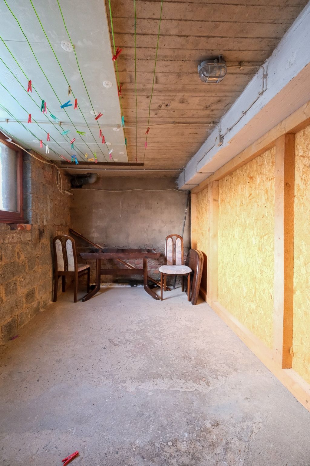A sparsely furnished room with a concrete floor, wooden chairs, and a table, clotheslines on the ceiling, and a partially open window side.