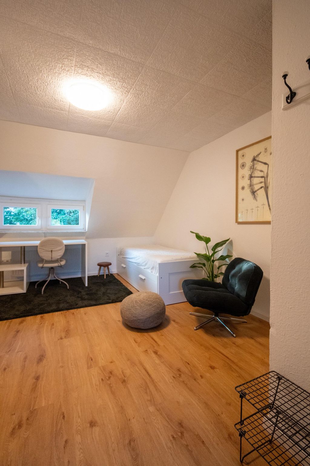 A minimalist room with light-colored walls, a white bed, a desk with a chair in front of a window, a black armchair next to a green plant, and a gray pouf on a wooden floor.