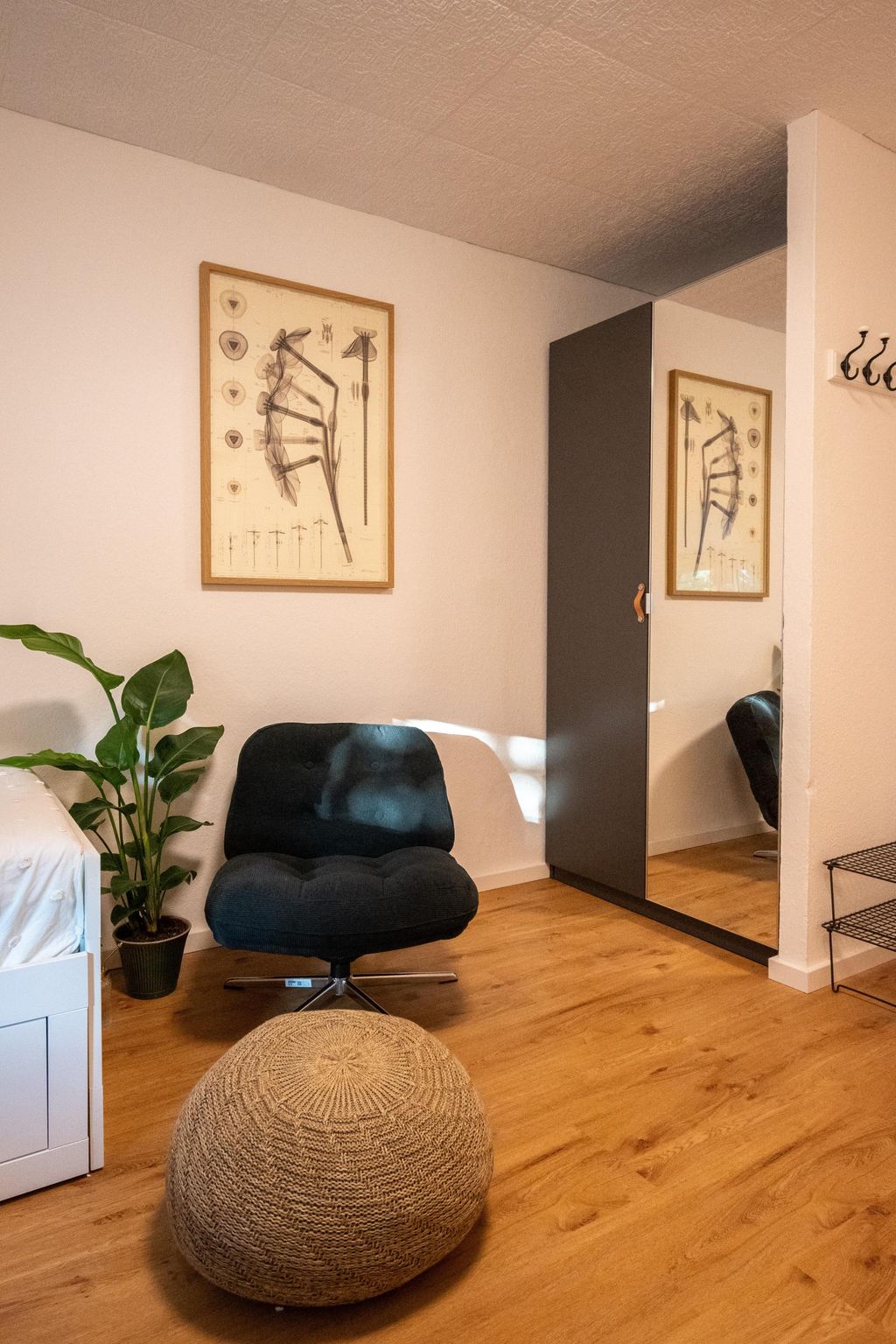 A cozy room featuring a dark blue armchair, a round knitted ottoman in front of a wardrobe with a mirrored door, a potted plant next to a white bed, and a framed botanical wall art.