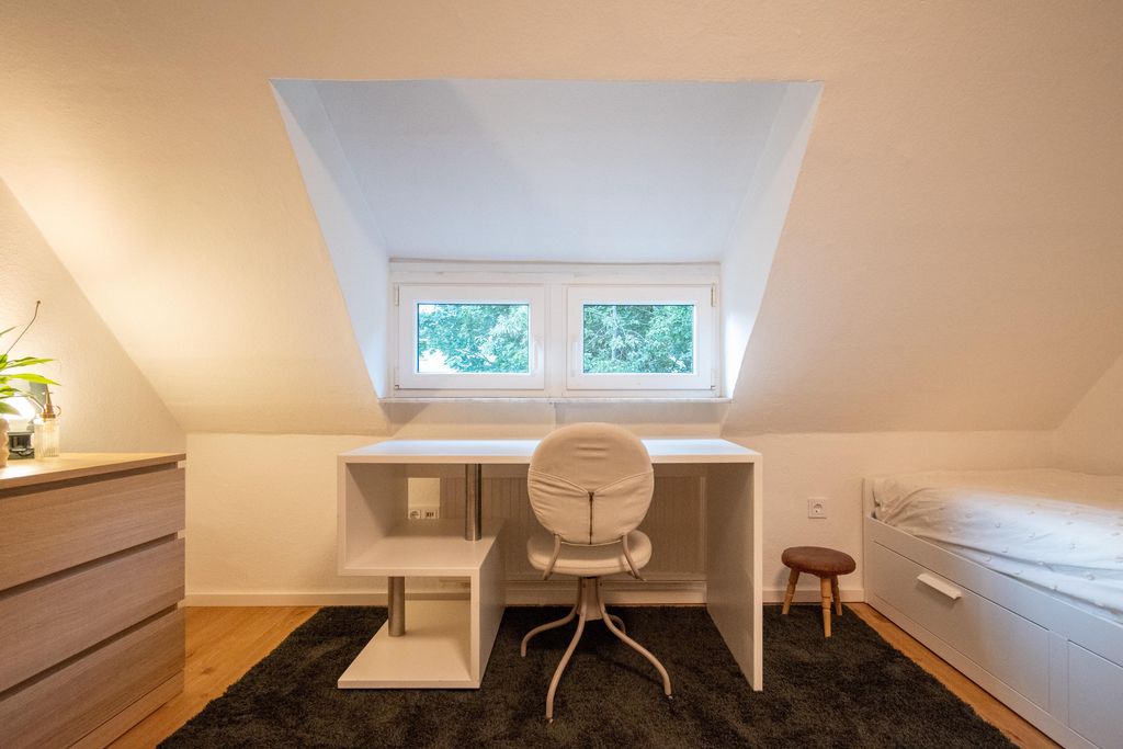 Bright minimalist room with a white desk in front of a dormer window, a gray office chair, a small stool, and a bed with white drawers under the slanted ceiling.