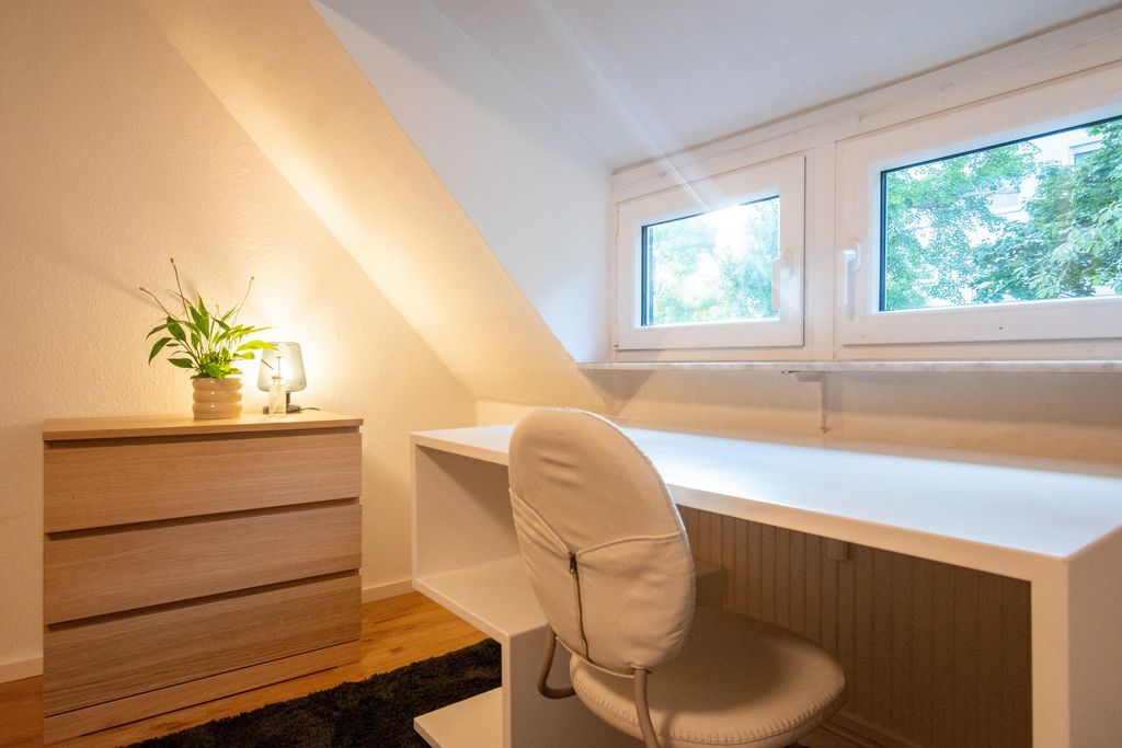 Bright room with a white desk, grey chair, and a small wooden dresser with a plant and lamp under a slanted ceiling.