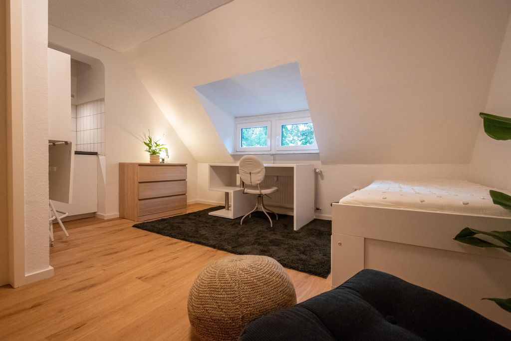 A simply furnished room with a sloped ceiling, a white desk in front of a small window, a white chair, a bed to the right, and a wooden dresser with a plant.